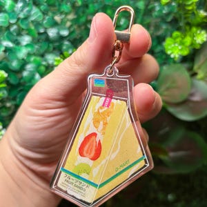 May include: A clear acrylic keychain featuring a fruit sando design. The keychain has a gold-colored clasp and a rectangular shape with rounded edges. The fruit sando illustration includes a strawberry, kiwi, and other fruits.