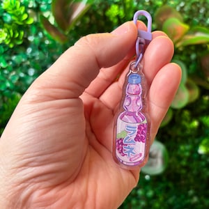 May include: A clear acrylic keychain featuring a purple soda bottle design with the Japanese text "Ramune Grape". The keychain has a purple clasp and a blue bottle cap. The background is blurred green foliage.