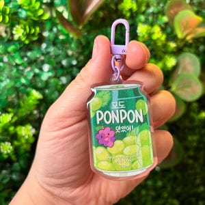 May include: A clear acrylic keychain featuring a green and white can design with the text "PONPON" and a purple grape illustration. The keychain has a purple clasp and a silver ring. The can design includes the text "fresh grape juice with pulp" and the measurement "255 ml".