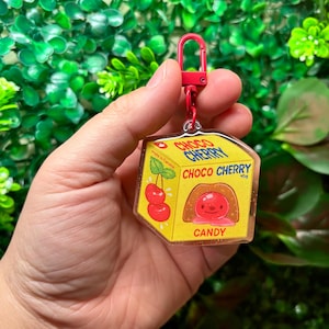 May include: A yellow Choco Cherry candy box keychain with a red clasp. The box features a cartoon cherry design and the words "Choco Cherry Candy". The keychain is held against a background of green foliage.