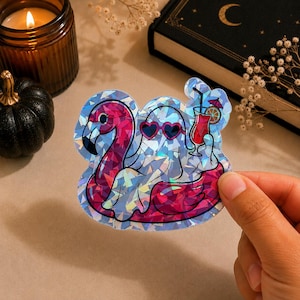 Op de afbeelding: Een holografische sticker met een witte geest die een roze hartvormige zonnebril draagt en in een felroze flamingo zwemband zit. De geest houdt een rode tropische cocktail vast met een klein parasolletje en een schijfje sinaasappel als garnering.