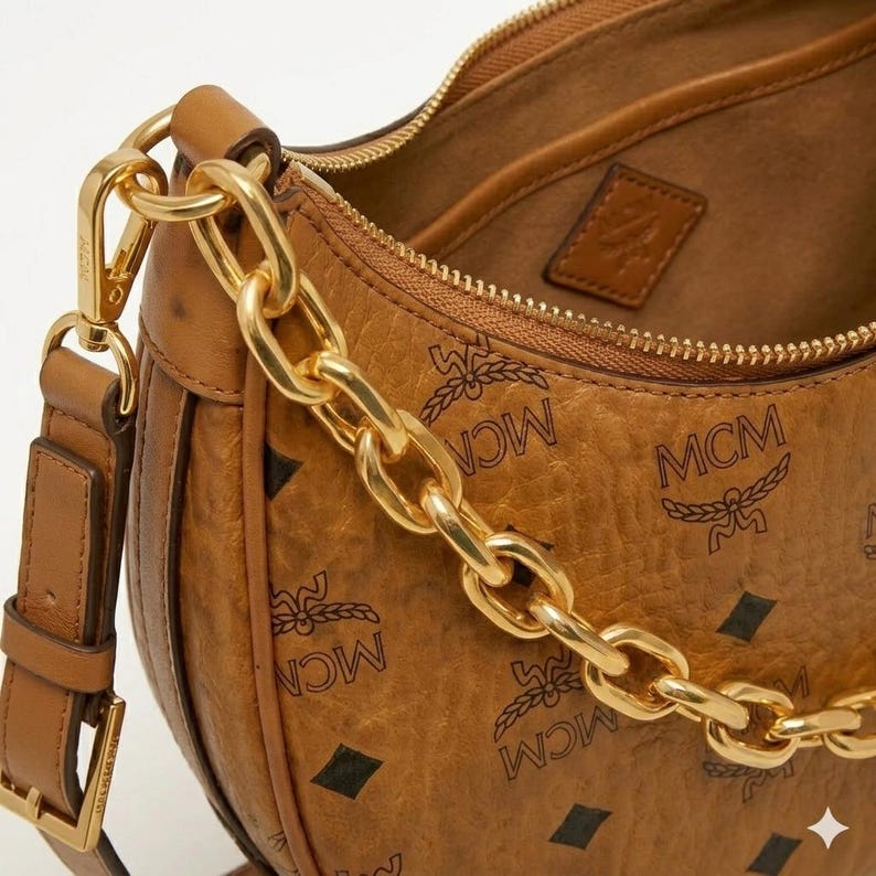 Pu&ograve; includere: Borsa a tracolla in pelle marrone con catena dorata. La borsa presenta un motivo ripetuto con il logo MCM e una cerniera dorata. L'interno della borsa &egrave; visibile, mostrando una fodera marrone e una piccola etichetta in pelle con un logo.