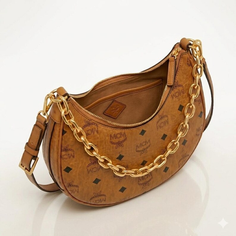 Pu&ograve; includere: Una borsa a spalla marrone con catena dorata e motivo con logo MCM. La borsa ha una forma curva, una chiusura con cerniera e una piccola tasca interna. La borsa &egrave; realizzata in materiale strutturato.