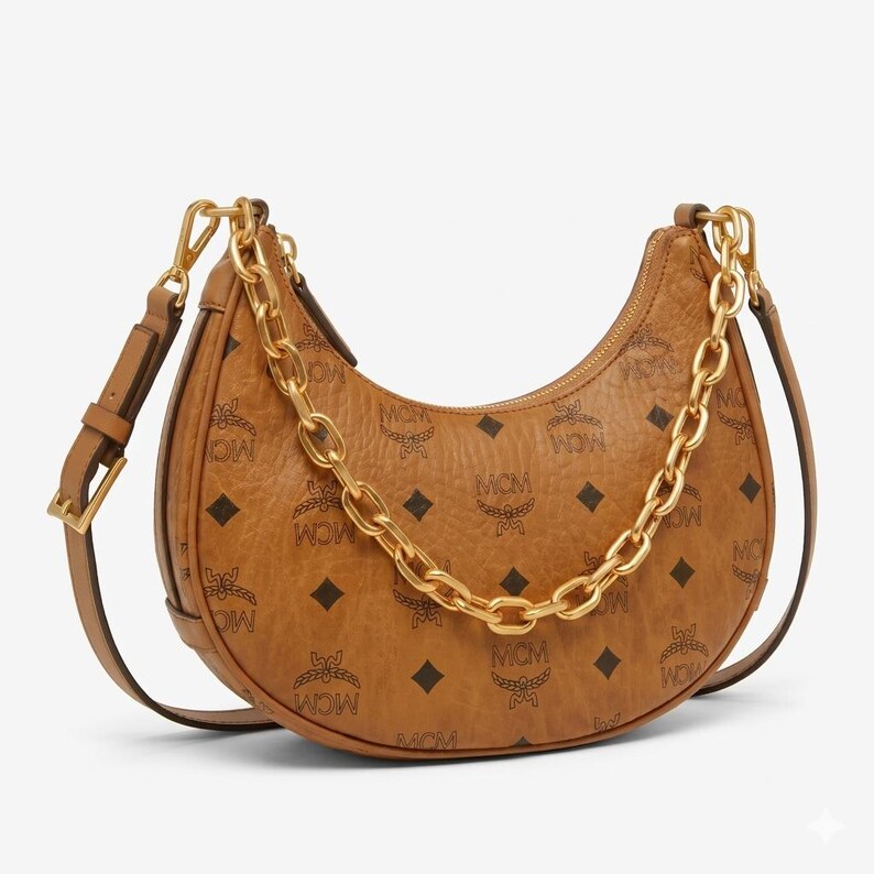 Pu&ograve; includere: Una borsa a tracolla in pelle marrone con una catena dorata e una tracolla regolabile abbinata. La borsa presenta un motivo ripetuto delle lettere "MCM" e forme a diamante. La borsa ha una forma curva e una chiusura a cerniera.