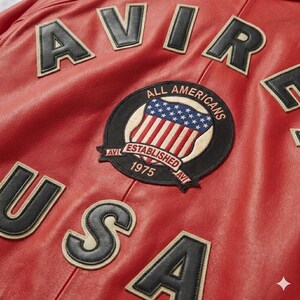 Peut inclure: Gros plan sur une veste en cuir rouge avec des lettres et des &eacute;cussons noirs et beiges. La veste porte les mots "AVIR" et "USA" en grandes lettres. Un &eacute;cusson circulaire indique "ALL AMERICANS ESTABLISHED 1975".