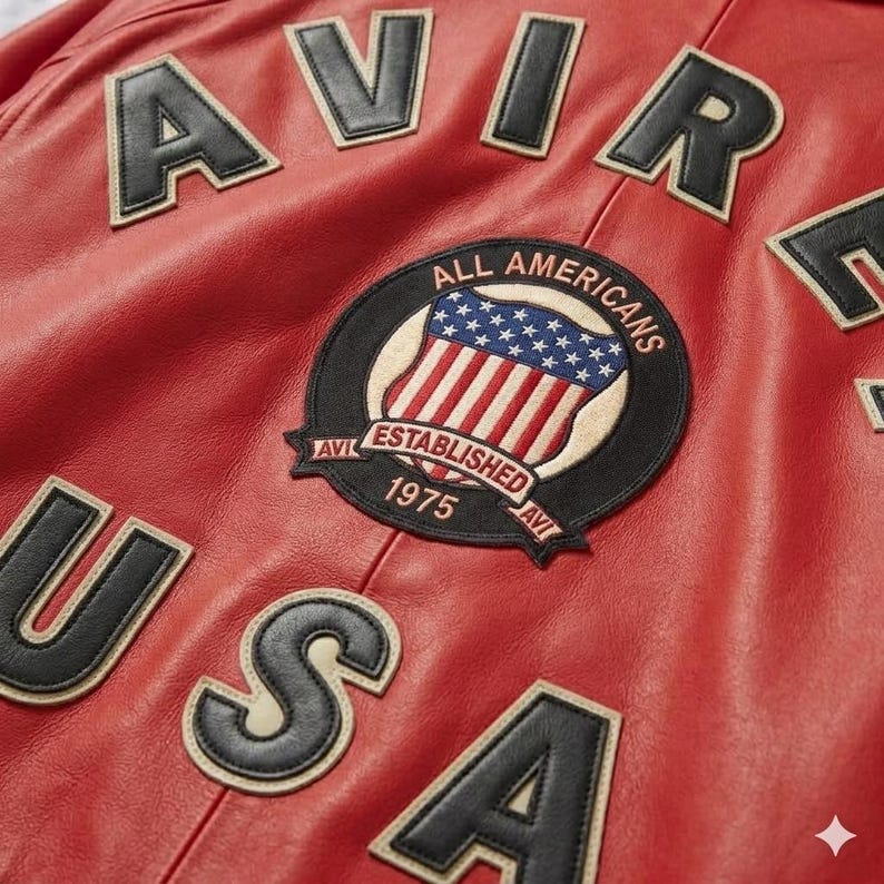 Peut inclure: Gros plan sur une veste en cuir rouge avec des lettres et des &eacute;cussons noirs et beiges. La veste porte les mots "AVIR" et "USA" en grandes lettres. Un &eacute;cusson circulaire indique "ALL AMERICANS ESTABLISHED 1975".
