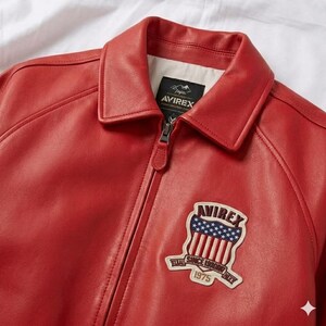 Peut inclure: Veste en cuir rouge avec un col et une fermeture &eacute;clair. La veste pr&eacute;sente un &eacute;cusson sur la poitrine avec la marque "AVIREX" et un motif de drapeau am&eacute;ricain. La veste a un style classique et vintage.