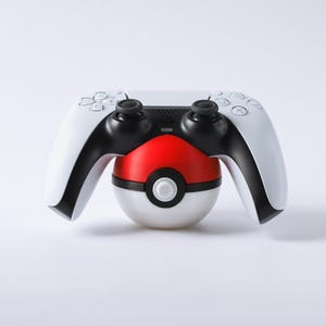 May include: A white and black PlayStation 5 controller resting on a red, white, and black Poké Ball stand. The controller has black joysticks and buttons. The stand is round with a white button in the center.