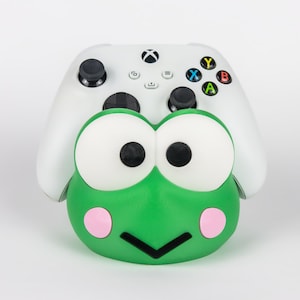 Peut inclure: Une manette de jeu vidéo blanche reposant sur un support en forme de grenouille verte. Le support présente de grands yeux blancs avec des pupilles noires, des joues circulaires roses et une bouche noire en forme de V. Manette avec joysticks noirs.