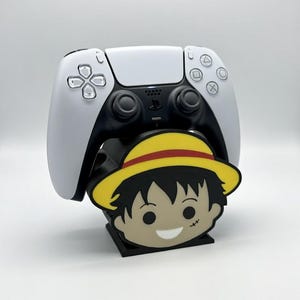May include: A white and black PlayStation 5 controller on a black stand with a cartoon character design. The stand has a yellow hat and smiling face. The stand is designed to hold the controller upright.