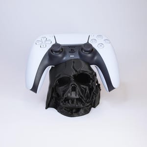 May include: A black Darth Vader helmet-shaped controller stand supporting a white PlayStation 5 controller. The controller has black accents and buttons. The stand is a unique accessory for gamers.