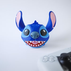 May include: A blue, cartoon-style character planter with large ears, a wide grin, and large eyes. The planter is a vibrant blue with pink accents inside the ears and mouth. A white game controller is in the foreground.