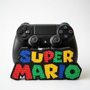 May include: Black PlayStation controller with a colorful "SUPER MARIO" logo. The logo features a knitted texture with red, yellow, blue, and green letters. The controller has buttons and joysticks.
