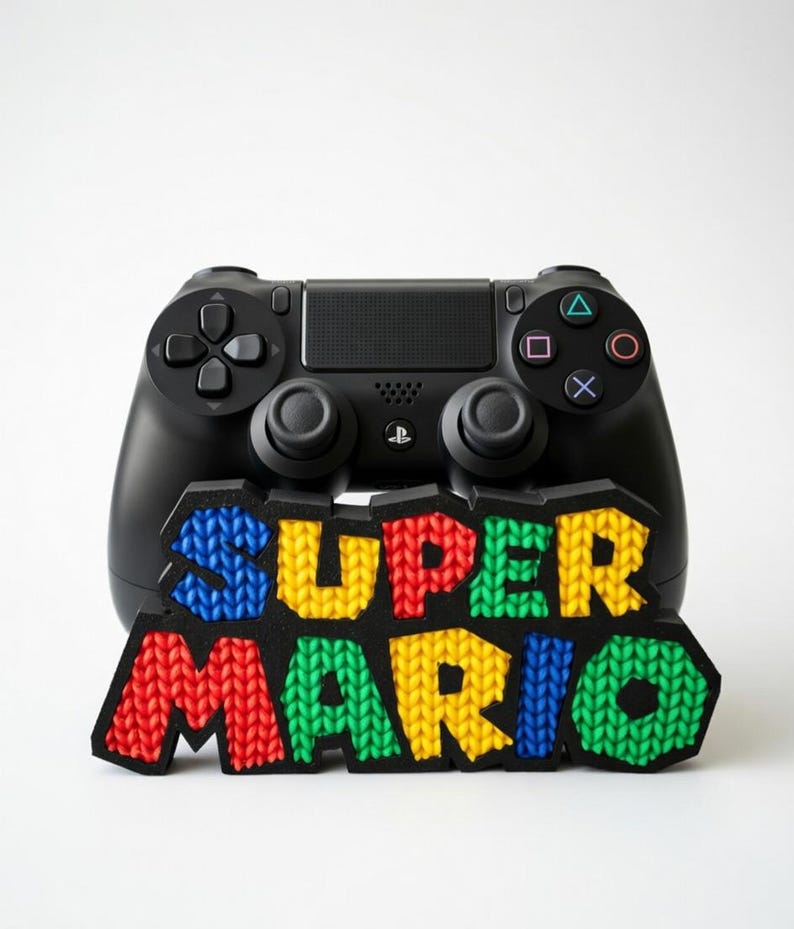 May include: Black PlayStation controller with a colorful "SUPER MARIO" logo. The logo features a knitted texture with red, yellow, blue, and green letters. The controller has buttons and joysticks.