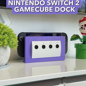 May include: A purple Nintendo Switch 2 GameCube dock with a white GameCube controller port design. The dock holds a black Nintendo Switch console. The text "NINTENDO SWITCH 2 GAMECUBE DOCK" is displayed in white on a purple background.