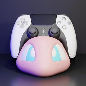 May include: A black and white video game controller resting behind a smooth, light pink character head featuring large, bright blue eyes. The controller includes a central PlayStation logo, dual analogue sticks, a directional pad, and four action buttons.