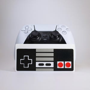 Op de afbeelding: Een witte en zwarte PlayStation 5-controller staat achter een gamecontroller in retrostijl. De retrocontroller is zwart-wit met rode knoppen, die doet denken aan een klassiek console-ontwerp.