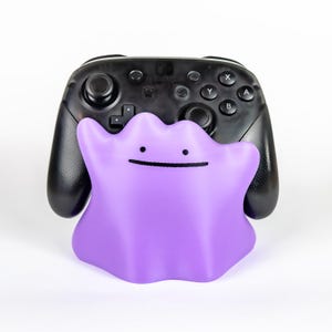 May include: A purple Ditto figure with a simple black facial expression is in front of a black Nintendo Switch Pro Controller. The controller has buttons and joysticks. The background is white.