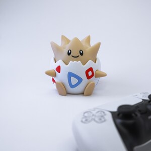 May include: A Togepi figurine emerging from a white and blue eggshell, with red and blue accents. The Togepi is tan with a smiling face. A white and black game controller is in the foreground.