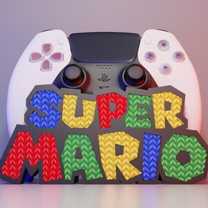 May include: A white and black game controller with the words "SUPER MARIO" in colorful, textured letters. The controller has pink and purple buttons. The background is a gradient of purple and red.
