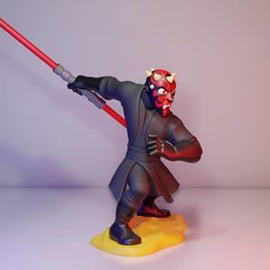 May include: A figurine of Darth Maul, a character from Star Wars, is depicted in a dynamic pose. The figure is predominantly black and red, holding a red lightsaber. The base is yellow. The character's face is detailed with red and black markings.