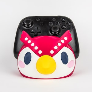 May include: A red, white, and yellow owl-shaped accessory sits in front of a black video game controller. The owl has large blue eyes, pink cheeks, and white dots on its head. The controller has buttons and joysticks.