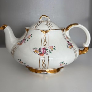 May include: White porcelain teapot with a lid, featuring gold accents on the handle, spout, and base. The teapot is decorated with floral patterns in pink, yellow, and green, and gold vertical lines.
