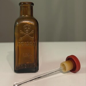 May include: An antique amber glass poison bottle, featuring a skull and crossbones symbol and the word "POISON". The bottle also has the text "LIC. IODINE". A dropper with a red cap and clear glass tube is next to the bottle.