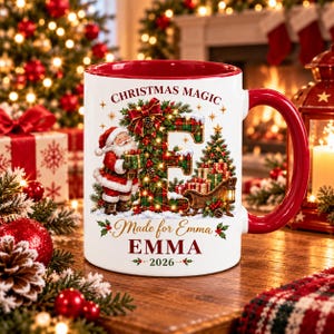 May include: A white ceramic mug with a red interior and handle, featuring a Christmas design with a plaid letter E, Santa Claus, a sleigh, and the text CHRISTMAS MAGIC, Made for Emma, EMMA, and 2026. The graphic includes holly and pine.