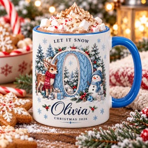 May include: A white ceramic mug with a blue handle and rim, filled with hot cocoa topped with whipped cream and marshmallows. The mug features a large blue letter O, a deer, a snowman, and the text LET IT SNOW, Olivia, and CHRISTMAS 2026.