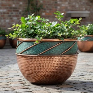 May include: A large, round copper planter with a hammered texture on the lower half. The upper section is a teal color with copper diagonal accents. Green plants spill over the rim, creating a striking contrast.