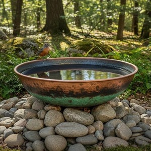 Puede incluir: Un bebedero de cobre con textura martillada, lleno de agua, descansa sobre un lecho de piedras. El cuenco tiene un borde oscuro y una pátina verde en la base. Un pequeño pájaro se posa en el borde, con un fondo de bosque.