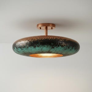 May include: A copper-colored ceiling light fixture with a hammered texture. The light has a circular, flattened shape with a dark green and black patina on the underside. The fixture is mounted on a copper-colored stem and base.