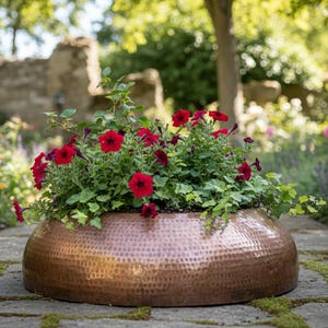 Op de afbeelding: Een koperen plantenbak met gehamerde afwerking, gevuld met levendige rode petunia's en groene bladeren, staat op een stenen pad. De komvorm van de plantenbak contrasteert met de tuinachtergrond en creëert een focuspunt.
