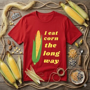 Op de afbeelding: Rood t-shirt met een afbeelding van een maïskolf en de gele tekst "I eat corn the long way". De afbeelding bevat maïskolven, korrels en decoratieve elementen op een houten oppervlak, wat een boerderijthema suggereert.