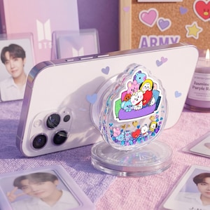 May include: A clear, decorative phone grip with a cartoon character design, attached to a white phone. The grip features a clear, rounded top filled with colorful confetti and character figures. The phone is surrounded by BTS-themed items.
