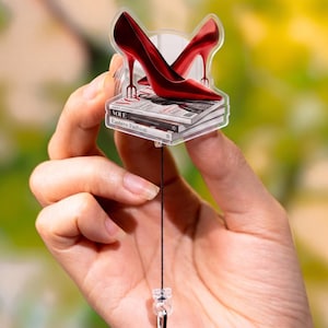 May include: A clear acrylic badge featuring a pair of glossy red high-heeled shoes atop a stack of magazines. The magazines have the word "Vogue" and "Eastern Fashion" printed on them. The badge is attached to a retractable reel with a clip.