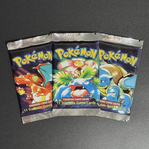 Può includere: Tre confezioni sigillate del gioco di carte collezionabili Pokémon. Ogni confezione presenta un'illustrazione di un personaggio Pokémon diverso, tra cui Charizard, Venusaur e Blastoise. Le confezioni sono argentate e blu con il logo Pokémon e il testo che indica che contengono 11 carte collezionabili.