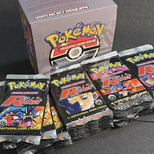 Può includere: Una collezione di bustine di carte collezionabili Pokémon e una scatola. Le bustine sono nere con grafiche colorate e la parola "Rocket". La scatola presenta il logo Pokémon e il testo "Team Rocket's on the Loose!"