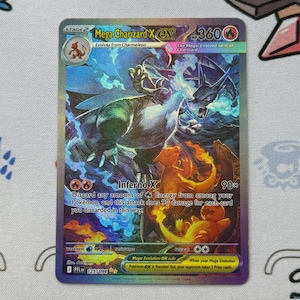 Puede incluir: Una carta coleccionable Pokémon Mega Charizard X EX holográfica. La carta presenta una ilustración detallada de una criatura parecida a Charizard con llamas y un fondo oscuro. El texto dice "Inferno X" y "Mega Evolution EX rule".