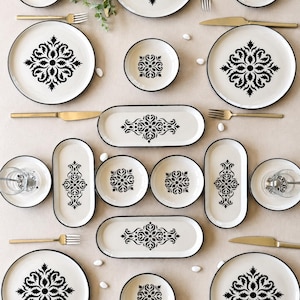 Può includere: Un set di stoviglie composto da piatti, ciotole e piatti da portata ovali color crema con motivi floreali neri decorati e bordi neri sottili. Il set include forchette e coltelli color oro disposti in modo simmetrico.