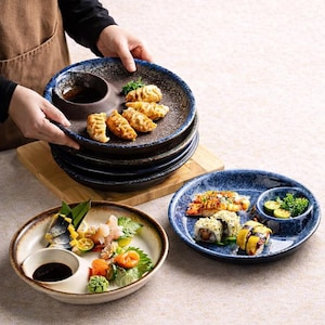May include: A stack of dark blue and brown ceramic plates, each with a built-in sauce dish. The plates are filled with dumplings. Two other plates with sushi and sashimi are also shown.