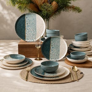May include: A dinnerware set featuring round plates and bowls with a reactive glaze finish in shades of deep blue, teal, and speckled cream. The collection includes stacked plates, nested bowls, and gold-toned cutlery arranged on a table.