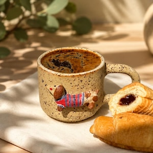 Puede incluir: Una taza de cerámica beige moteada llena de café oscuro, con un perro salchicha esculpido en tres dimensiones que lleva un suéter de rayas rojas y azules. La taza tiene un asa curva y un acabado texturizado y moteado en toda su superficie.