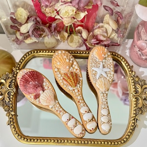 May include: Three wooden hairbrushes with seashell and pearl embellishments rest on a gold-framed mirror. The brushes feature various seashell designs in shades of pink, orange, and white, with pearl accents. The mirror is on a white surface.