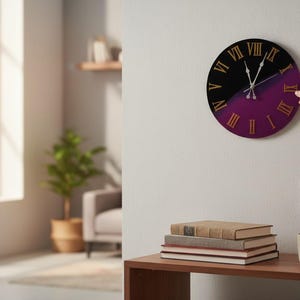 May include: A round wall clock with a black and purple design. The clock has gold Roman numerals and hands. A person's hand is adjusting the clock. The clock is on a white wall. A wooden table holds books and a mug. The clock's diameter is approximately 30 cm.