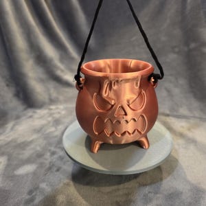 May include: A metallic copper-colored cauldron with a sculpted, angry jack-o'-lantern face featuring sharp eyes and a jagged mouth. The round vessel stands on three small feet and includes a thin black cord handle attached to the sides.