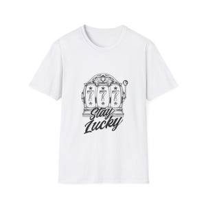 以下が含まれることがあります： 白のTシャツ。前面に、数字の777と「Stay Lucky」の文字が入った、白黒のスロットマシンのグラフィックがプリントされています。クルーネックと半袖のデザインです。