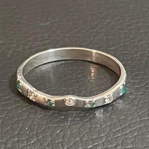 May include: A silver ring with a wavy design, set with small emerald and clear stones. The ring has a polished finish and is displayed on a dark surface. The ring is a piece of jewellery.