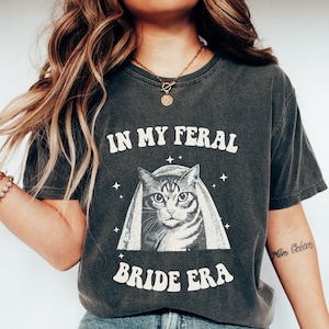 May include: Dark gray t-shirt with the text "IN MY FERAL BRIDE ERA" and a graphic of a cat wearing a veil. The cat graphic is in black and white. The shirt is a vintage style.